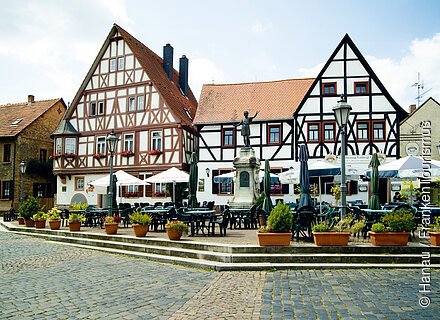 Steinheim - Platz des Friedens Marktplatz mit Fachwerkhäusern, Sitzgelegenheiten, Sonnenschirmen und einer Statue in der Mitte